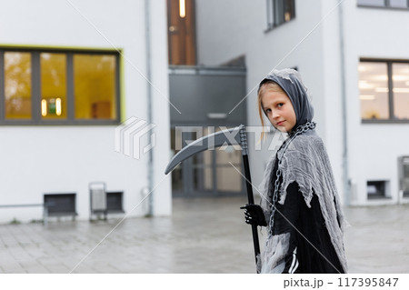Portrait of kid in grim reaper Halloween costume with scythe and bag for sweets at school building facade ready for Halloween party celebration. Child in scary spooky black death suit outdoors 117395847