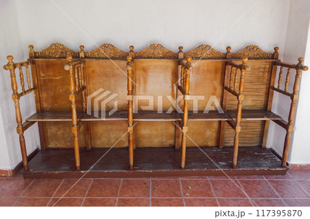 A set of empty, vintage wooden church chairs with intricate carvings arranged outdoors against a white wall and floor with reddish-brown tiles. A set of empty, vintage wooden church chairs with intricate carvings arranged outdoors against a white wall and floor with reddish-brown tiles. 117395870