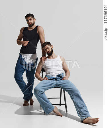 Two men in jeans and tank tops strike relaxed pose, one standing confidently while other leans on chair, exuding casual, stylish vibe against warm grey background. 117396194