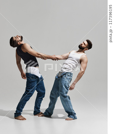 Two men in casual jeans and tank tops lean back, holding hands, creating dynamic pose that balances strength and connection against warm grey background. 117396195