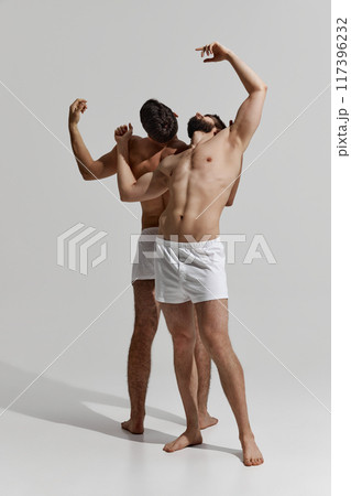 Two men in white underwear strike expressive poses, their bodies intertwined in moment of fluid motion against warm grey background. Two men in white underwear strike expressive poses, their bodies intertwined in moment of fluid motion against warm grey background. 117396232