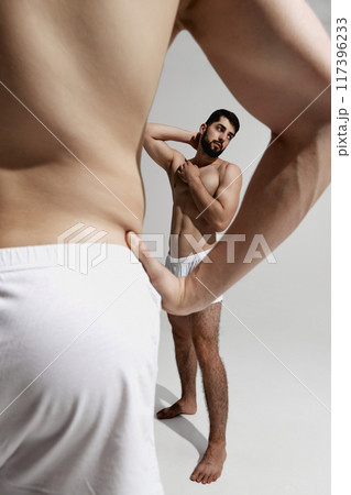 Thoughtful man in white underwear poses with one hand behind his head, framed by blurred silhouette of another. 117396233