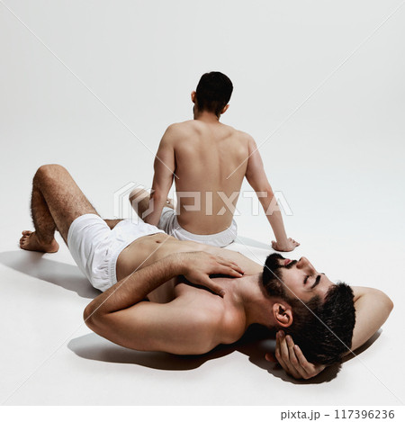 Two men in white shorts relax, one lying down with hand on his chest, other sitting upright, both reflecting moment of calm and rest. 117396236