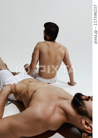 Two men in white shorts relax, one lying down with hand on his chest, other sitting upright, both reflecting moment of calm and rest. Two men in white shorts relax, one lying down with hand on his chest, other sitting upright, both reflecting moment of calm and rest. 117396237