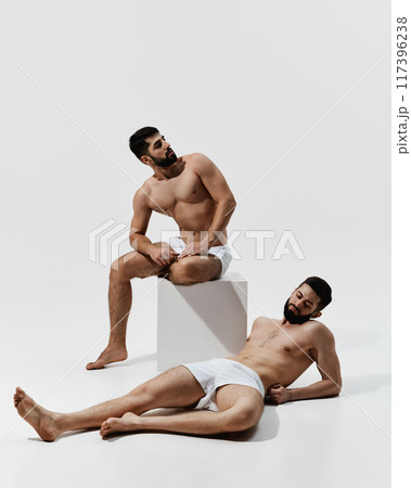 Two men in white underwear rest, one seated on white block, other reclining on floor against warm grey background. Two men in white underwear rest, one seated on white block, other reclining on floor against warm grey background. 117396238
