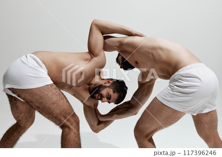 Two men, athletes grapple, showcasing their strength and technique against warm grey studio background. 117396246