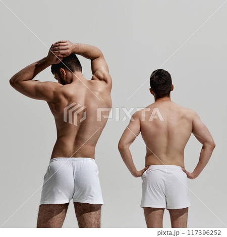 Rear view photo of two young men posing shirtless showing their healthy backs and postures against warm grey background. 117396252