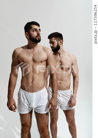 Two brutal male models with beards, with muscular build bodies posing shirtless, in pants against warm grey background. Two brutal male models with beards, with muscular build bodies posing shirtless, in pants against warm grey background. 117396254