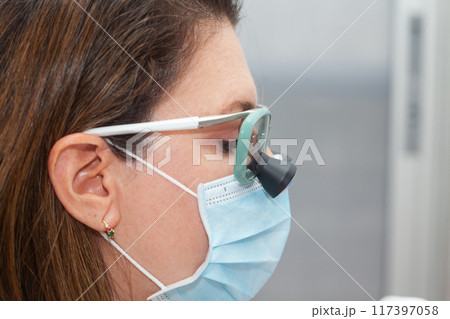 Close up of a female dermatologist face while suturing a patient during a small surgery wearing a binocular loupe. Close up of a female dermatologist face while suturing a patient during a small surgery wearing a binocular loupe. 117397058