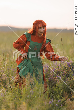 A shepherdess woman with red hair and a green dress sits in a field of flowers. 117398110