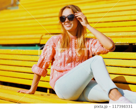 Portrait of happy smiling young blonde woman sitting on bench in a city park Portrait of happy smiling young blonde woman sitting on bench in a city park 117398331