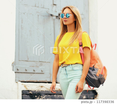 Stylish blonde young woman in summer yellow t-shirt with backpack, glasses posing in the city 117398336