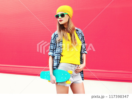 Stylish cool blonde woman with skateboard wearing colorful yellow hat on pink background Stylish cool blonde woman with skateboard wearing colorful yellow hat on pink background 117398347