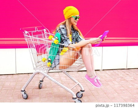 Stylish young woman sitting in trolley with tablet pc and skateboard over colorful pink background Stylish young woman sitting in trolley with tablet pc and skateboard over colorful pink background 117398364