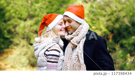 Winter portrait of happy smiling young couple hugging in red santa hats outdoors, christmas tree 117398434