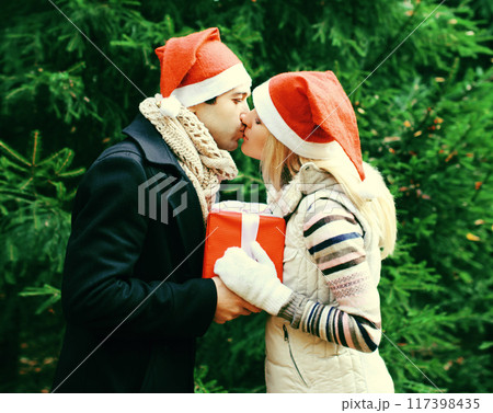 Winter portrait of happy young couple kissing in santa hats with gift box outdoors, christmas tree Winter portrait of happy young couple kissing in santa hats with gift box outdoors, christmas tree 117398435