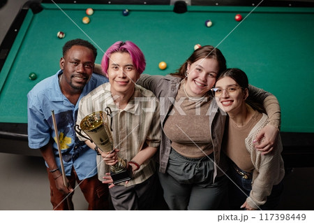 Group of friends standing by pool table, holding trophy and smiling in celebration of recent victory, basking in moment of joy and camaraderie 117398449
