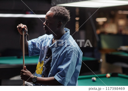 Man focusing on making shot while playing billiards in dimly-lit pool hall. Holding cue stick with concentration, adjusting stance for precision without looking at camera 117398469