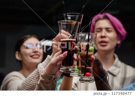 Three friends enjoying drinks at gathering while raising glasses in toast. Smiling and showing camaraderie, each holding different beverage in lively social setting 117398477