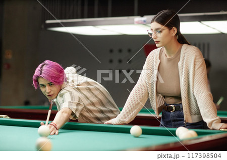 Young man with pink hair aiming billiard cue while woman with glasses watching attentively in billiard hall having some focus 117398504