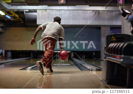 Black person enjoying evening by bowling down lane while wearing casual attire holding shining red ball aimed toward pins Black person enjoying evening by bowling down lane while wearing casual attire holding shining red ball aimed toward pins 117398531