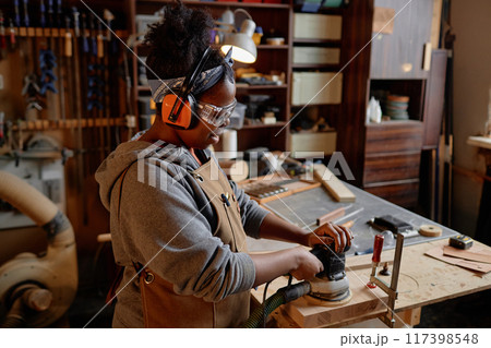 Young Black woman in workshop industriously sanding wood with protective gear surrounded by woodworking tools and materials Young Black woman in workshop industriously sanding wood with protective gear surrounded by woodworking tools and materials 117398548