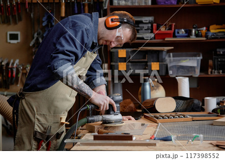 Artisan focusing on sanding wooden piece using electric sander in well-equipped woodworking workshop with various tools and materials arranged on shelves 117398552