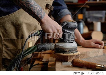 Detailed image showing craftsman using electric sander on wooden piece in workshop. Forearm with tattoo visible, emphasizing intricate work on smoothening wood surface 117398553