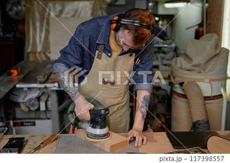 Person seen concentrating on sanding wood piece with sander machine in workshop filled with various tools and gadgets surrounding. Wearing protective gear and focused on task 117398557