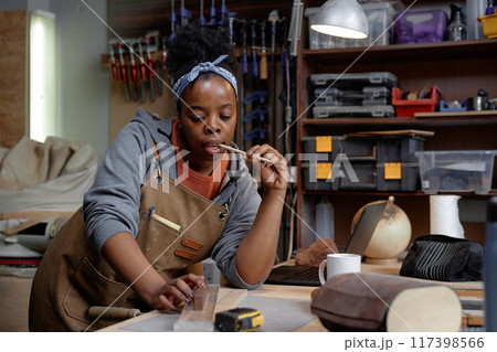 Woman wearing apron carefully carving wood in her workshop surrounded by tools and equipment focused on creating detailed piece of art 117398566