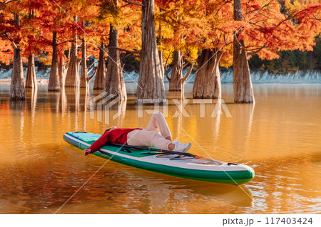 Traveller woman lies on stand up paddle board at the lake between trees in autumnal season with morning sunlight 117403424