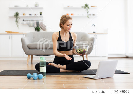 Woman practicing yoga at home with healthy salad and laptop Woman practicing yoga at home with healthy salad and laptop 117409007