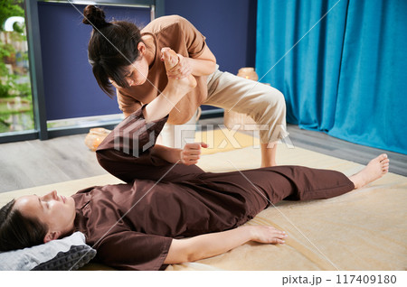 Masseuse making thai yoga massage. Woman therapist doing traditional massage treatment for female patient indoors. Client lying on mat, meditating, enjoying hand massage in calm atmosphere. 117409180