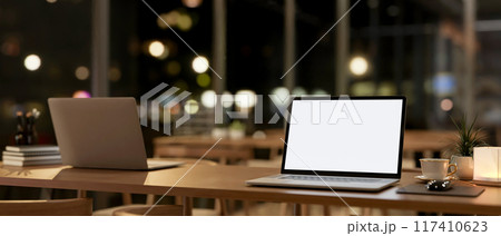 A laptop computer mockup on a wooden table in a contemporary coffee shop co-working space at night. 117410623