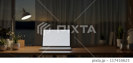 A laptop computer on a wooden table by the window in a house at night, illuminated by a table lamp. A laptop computer on a wooden table by the window in a house at night, illuminated by a table lamp. 117410625