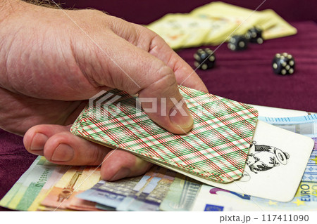 Man's hand holding a card over banknotes. The men are playing cards his hand. One joker card is opened. 117411090