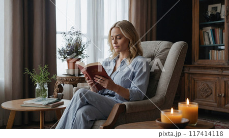 A woman sits comfortably in an armchair, reading a book in a cozy living room with soft lighting and candles on a table. A woman sits comfortably in an armchair, reading a book in a cozy living room with soft lighting and candles on a table. 117414118