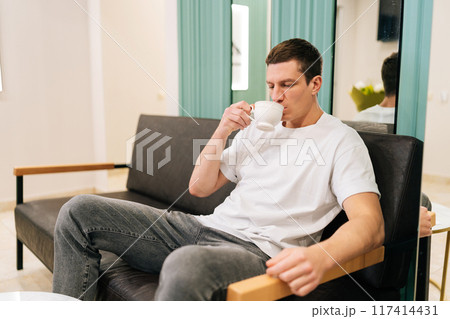 Successful male client sipping from cup of coffee while comfortably waiting in modern and welcoming lobby of medical clinic. Good looking male client enjoying cup of tea while relaxing in waiting room 117414431