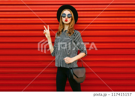 Stylish woman model wearing a black white striped shirt, hat and handbag clutch on red background Stylish woman model wearing a black white striped shirt, hat and handbag clutch on red background 117415157