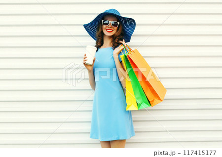 happy smiling young woman with coffee cup and shopping bags in blue dress, summer hat on white 117415177