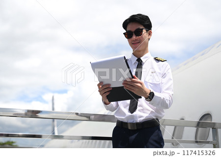 Pilot wearing sunglasses reading paper while standing beside an airplane with cloudy sky on background Pilot wearing sunglasses reading paper while standing beside an airplane with cloudy sky on background 117415326