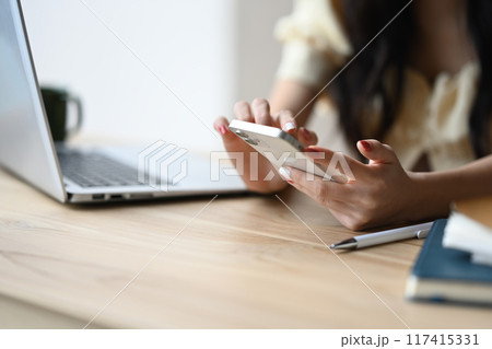 Close up shot of woman holding a cell phone in her hand while sitting at a desk with a laptop Close up shot of woman holding a cell phone in her hand while sitting at a desk with a laptop 117415331