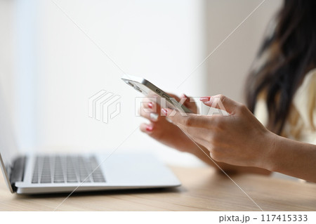 Young woman is holding a cell phone in her hand while sitting at a desk with a laptop 117415333