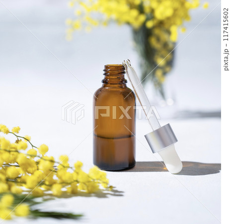 Face and body serum in bottle on a light background with mimosa flowers and hard shadow close up. Face and body serum in bottle on a light background with mimosa flowers and hard shadow close up. 117415602