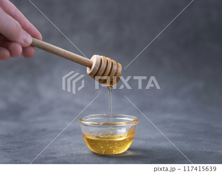 Honey flows from a wooden stick into a glass bowl on a dark background close-up. Honey flows from a wooden stick into a glass bowl on a dark background close-up. 117415639
