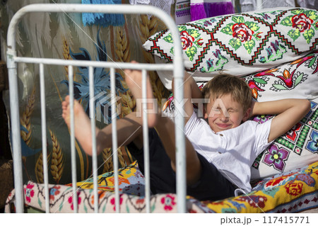 Child Russian or Ukrainian on an old ethnic bed with pillows and bedspreads embroidered with a Slavic pattern. Belarusian boy. 117415771