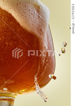 Close up shot of glass in condensation droplets filled with golden colored, bubbled and foamy beer against light background. Close up shot of glass in condensation droplets filled with golden colored, bubbled and foamy beer against light background. 117415821