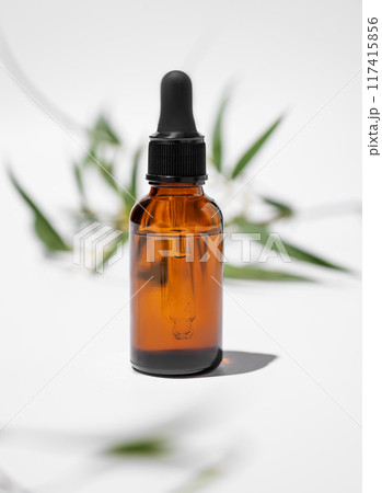 Face and body serum in bottle on a white background with green leaves and shadow close up. 117415856