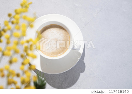 Cappuccino or latte with milk foam in a white cup on a light background with yellow mimosa flowers. 117416140