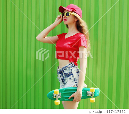 Young woman with green skateboard wearing a shorts and red baseball cap in a city 117417552